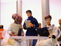 three action figures sitting at a table with a book in their hands and one on the other side