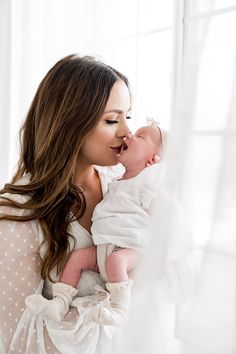 a woman holding a baby in her arms and kissing it's face with the window behind her