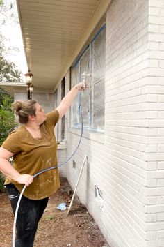 Woman using Graco Magnum 262800 X5 Stand Airless Paint Sprayer to paint exterior brick home white with paint sprayer. Instructions of how to paint a brick house with pros and cons. Blue painters tape with sheeting covering windows.