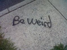 the word be weird written in black ink on concrete next to grass and street corner