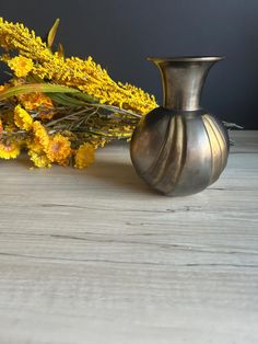 a silver vase sitting on top of a wooden table next to yellow wildflowers