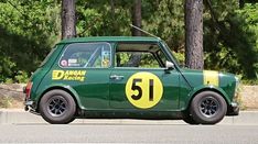 an old green car with yellow numbers parked on the side of the road in front of some trees