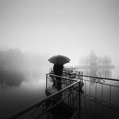 a person with an umbrella standing on a dock in the foggy water near a body of water