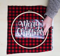 a person is stitching a merry christmas sign on a piece of red and black plaid fabric