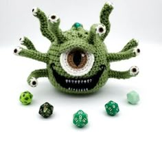 a green crocheted toy with eyes and teeth surrounded by dices on a white surface