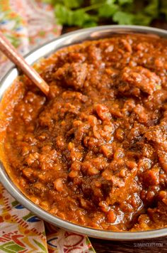 a bowl filled with chili and meat on top of a colorful table cloth next to a wooden spoon