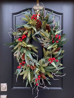 a christmas wreath hanging on the front door with holly and red berries, greenery and pine cones