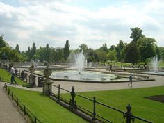 people are walking around in the park with water fountains on either side and green grass