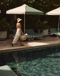 a woman in a white dress and straw hat walking by an outdoor pool with sun umbrellas