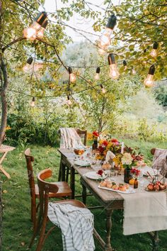 an outdoor dinner table set for two with lights hanging from the trees and flowers in vases