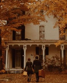 two people walking in front of a white house with autumn leaves on the ground and trees