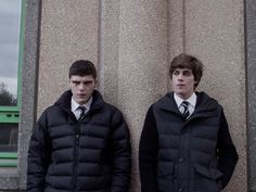 two young men standing next to each other in front of a wall wearing jackets and ties