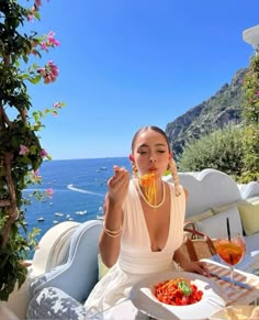 a woman sitting at an outdoor table with food and drinks in front of the ocean