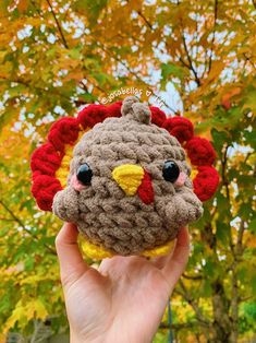 a hand holding a small crocheted chicken in front of a tree with leaves