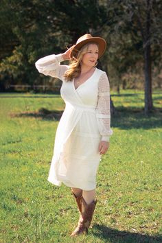 a woman in a white dress and hat is standing in the grass with her hands on her head