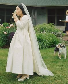 a woman in a wedding dress standing on the grass with a dog and holding a flower