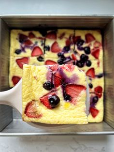 a square cake with berries and blueberries on it in a metal pan, being held by a spatula