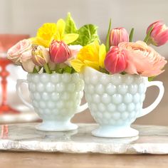 two white cups with flowers in them sitting on a table next to a vase filled with pink and yellow flowers