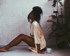 a woman sitting on the ground next to a plant and a coffee cup in front of her