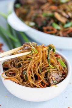 a white bowl filled with noodles and meat on top of a blue tablecloth next to another bowl full of vegetables