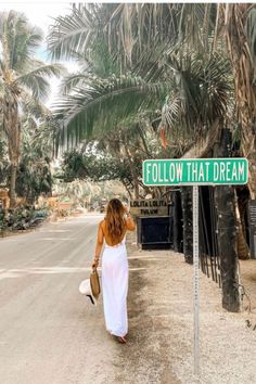 a woman in white dress walking down street next to palm trees and sign that says follow that dream