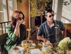 a man and woman sitting at a table with food in front of them, drinking coffee