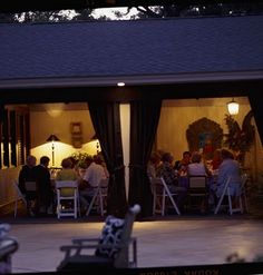 people sitting at tables in front of a house with lights on and patio furniture set up