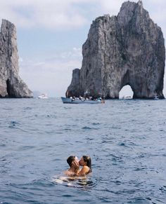 two people are kissing in the water near some cliffs and boats on the ocean behind them