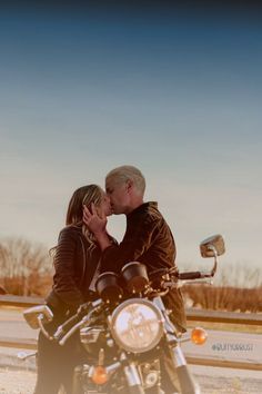 a man and woman kissing on the back of a motorcycle in front of a road