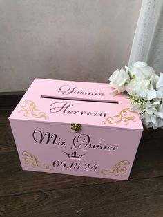 a pink box with gold lettering and flowers on the top is sitting next to a window