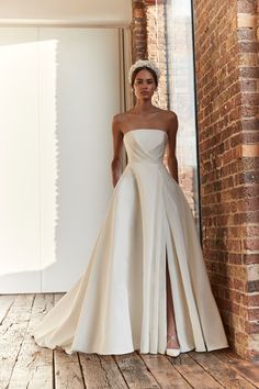 a woman standing in front of a brick wall wearing a wedding dress with high slit