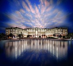 a large building sitting on top of a lake next to a lush green forest under a blue cloudy sky