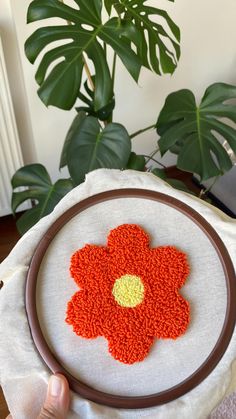 a person holding a cross - stitch flower on a white cloth in front of a potted plant