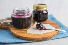 two jars of jam and a spoon on a cutting board with blue cloth next to them