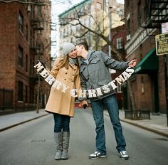 a man and woman kissing on the street while holding letters that say merry christmas in front of them