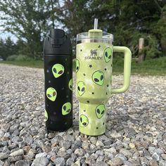 two travel mugs sitting next to each other on the ground with rocks and trees in the background