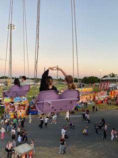 two people sitting on swings in the middle of an amusement park while others walk around