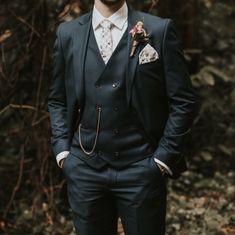 a man wearing a suit and tie standing in front of some trees with his hands in his pockets