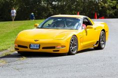 a yellow sports car driving down a road with people in the back ground looking at it