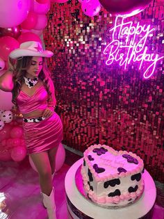 a woman standing next to a birthday cake with pink frosting and balloons in the background