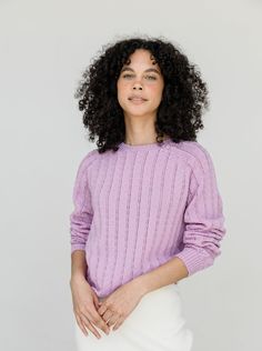 a woman with curly hair wearing a purple sweater and white skirt, posing for the camera