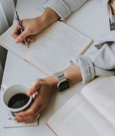 a person sitting at a table writing in a book and holding a cup of coffee