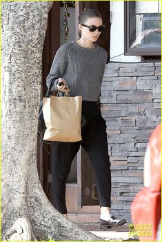 a woman in grey sweater and black pants carrying shopping bags while standing next to a tree