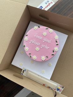 a pink cake with daisies on it in a cardboard box next to a pen