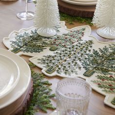 the table is set for christmas dinner with pine trees on it and white dishes in front