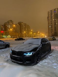 two cars are parked in the snow near some high rise buildings and street lights at night