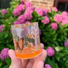 a hand holding an orange glass with a butterfly painted on it in front of pink flowers