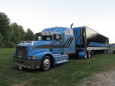 a large blue semi truck parked on top of a lush green field next to trees