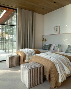 two beds in a room with large windows next to each other and foot stools on the floor