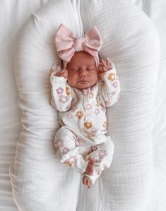 a baby is laying on a pillow with a pink bow in her hair and wearing a white outfit